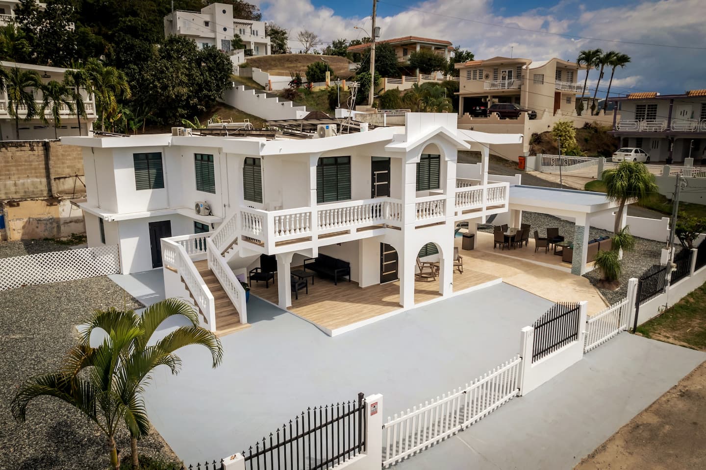 Aerial view of the white duplex villa with wraparound balconies, exterior staircase, covered terrace and gated driveway in a hillside Rincón neighborhood