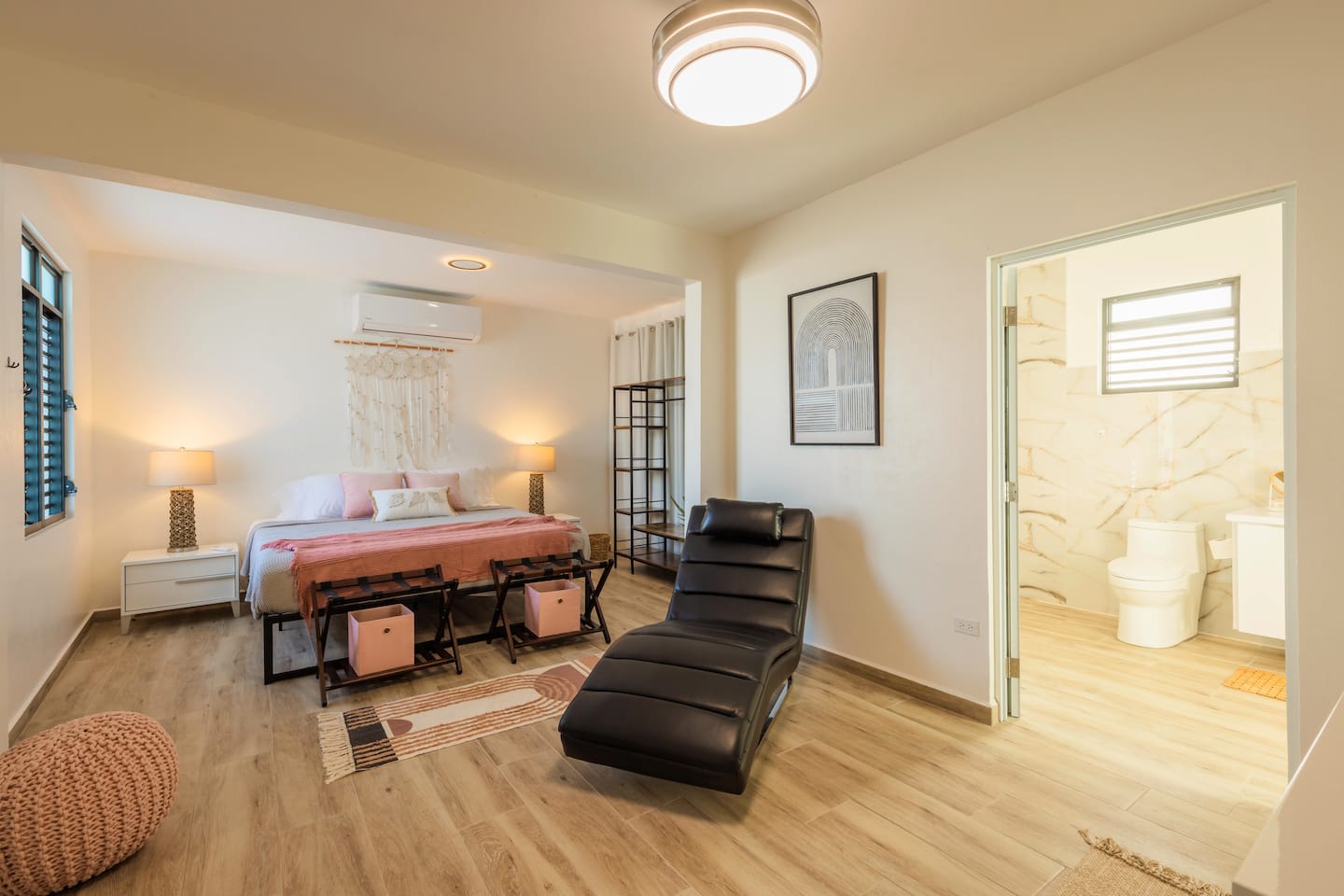 Master bedroom with a king bed, blush and pink accents, black leather chaise and an ensuite bathroom through the open door