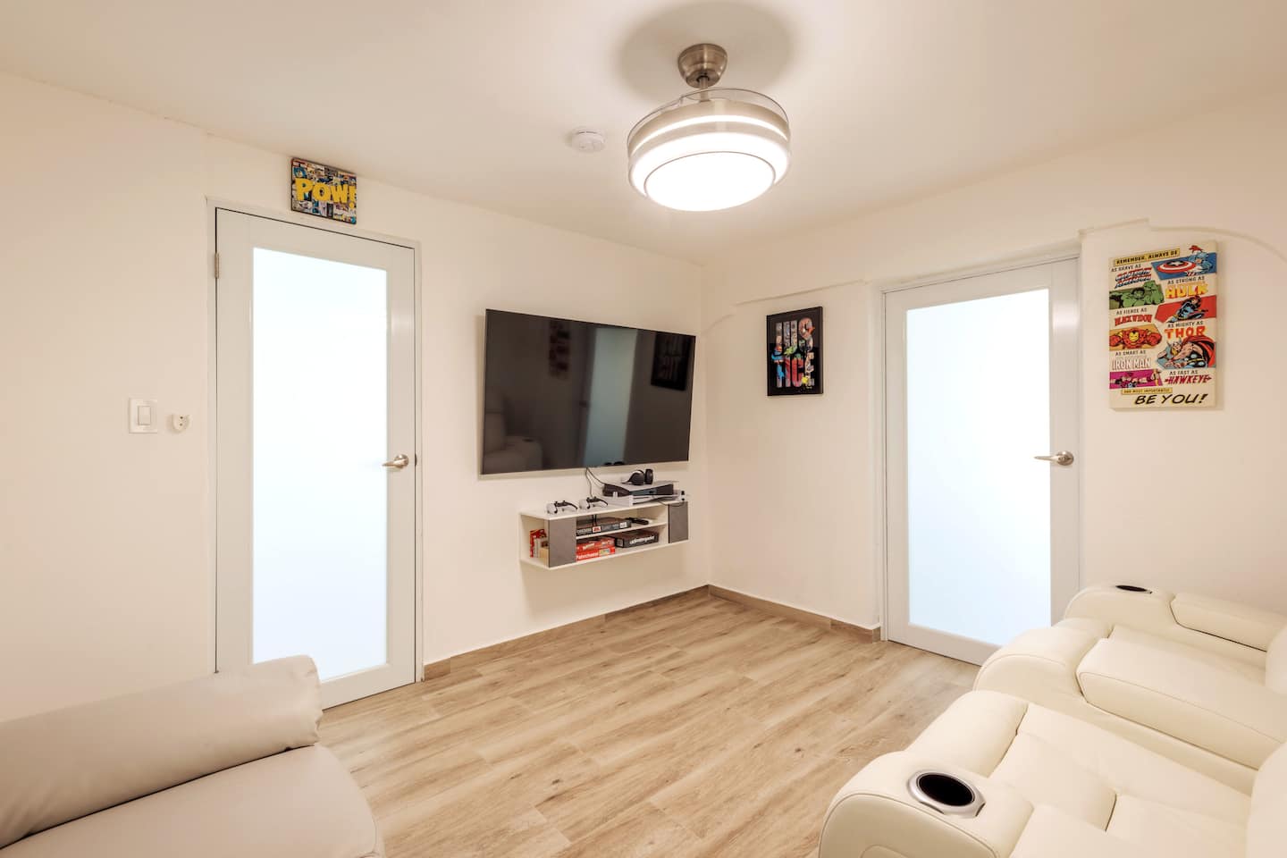 Game room with a wall-mounted TV over a console shelf, comic-book art on the walls and white recliner sofas