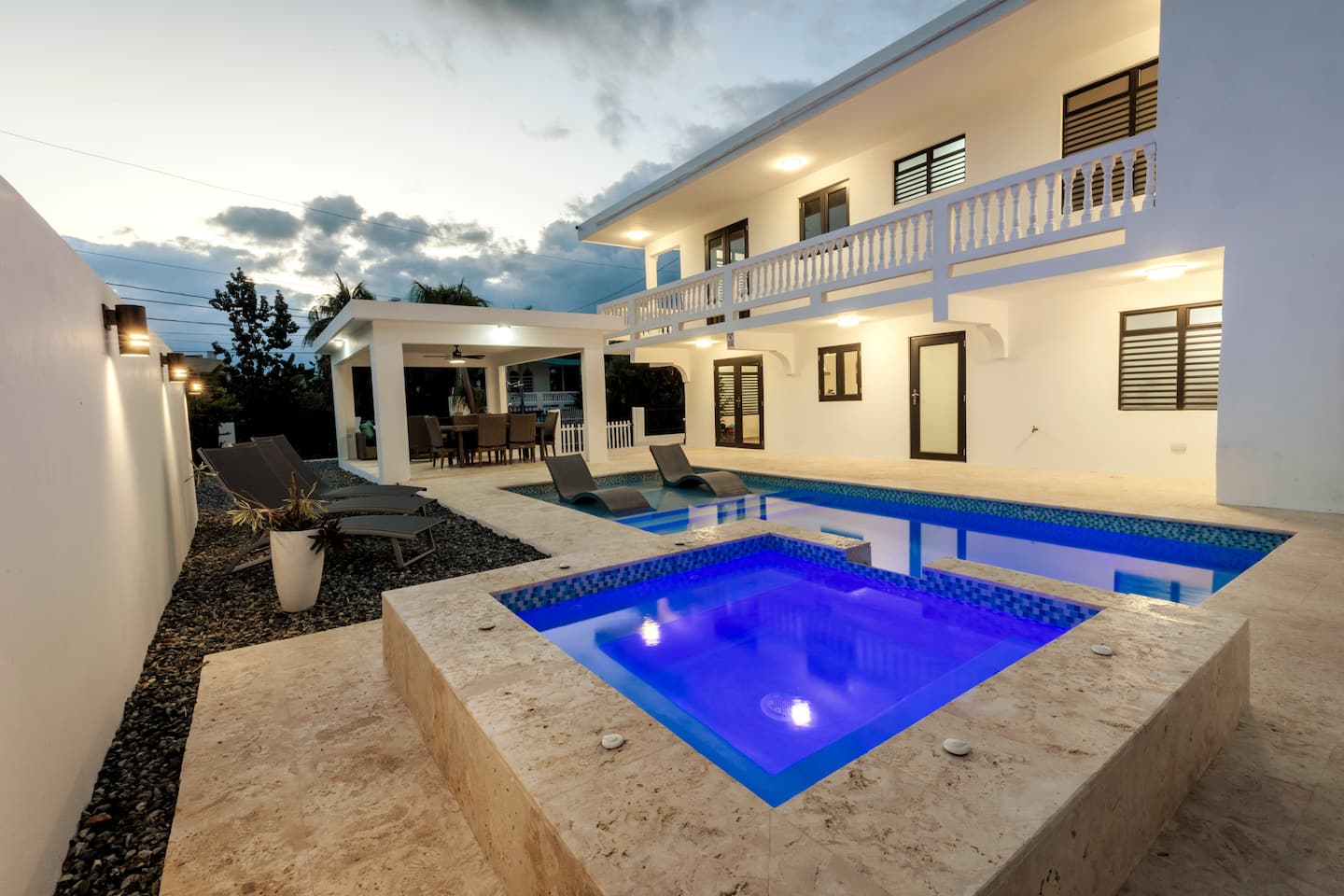 Casa Caleta at dusk: the heated pool and glowing blue jacuzzi in front of the white two-story villa, with the covered outdoor dining area lit at left