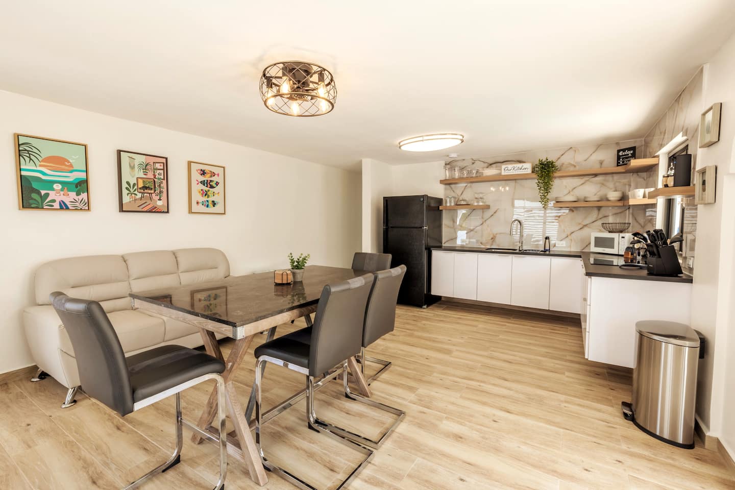 Open-plan living and dining room with a full kitchen, cream leather sofa, stone-top dining table and tropical art on the walls