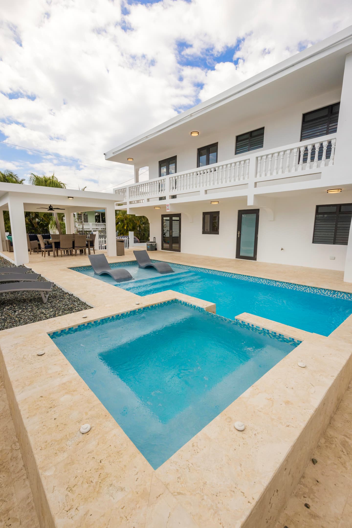 Daytime view of the pool and raised square jacuzzi with in-pool loungers, below the villa's white balustraded balcony
